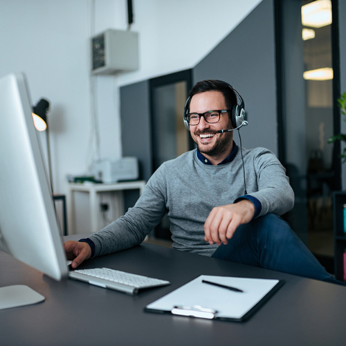 Smiling man wearing glasses and headset sitting at desk, looking at computer monitor in modern office.
